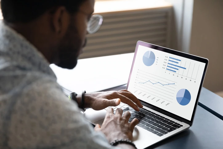 Focused young African ethnicity male marketing specialist professional working on project report, analyzing graphs charts in economic application on computer, sitting at table in office or home.