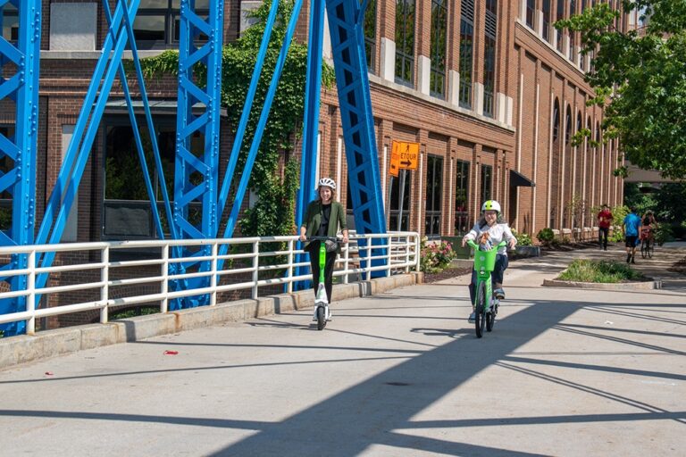 An adult riding a scooter and another riding a bike across a bridge