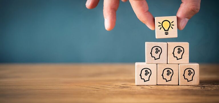 Small stacked wooden blocks each with a silhouette of a man's head, and a hand placing a top block of a lit light bulb representing an idea
