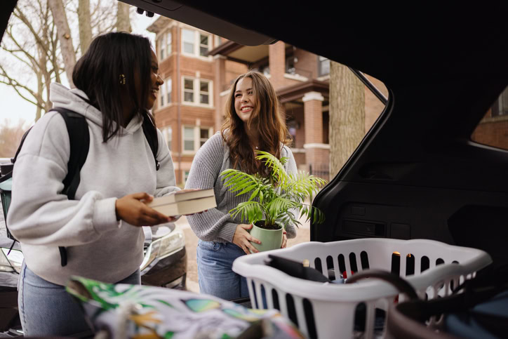 Two college students unloading belongings from a car trunk, excitedly moving into a new apartment and starting their independent lives