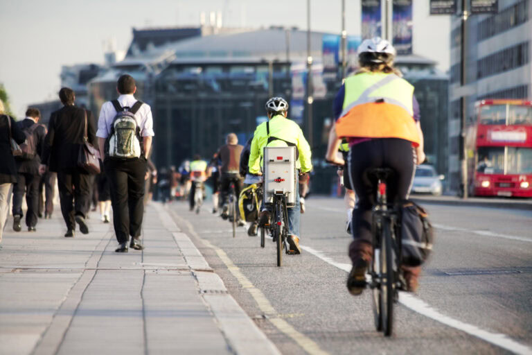 Commuters on the sidewalk, bikers on the biking lane, double decker bus in the background