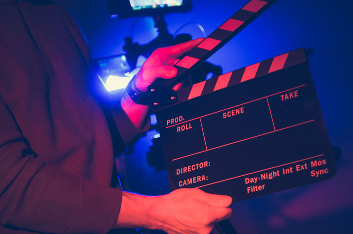 A filmmaker holds a clapperboard labeled with production details, preparing for a night shoot in vibrant lighting, showcasing the behind-the-scenes activity.