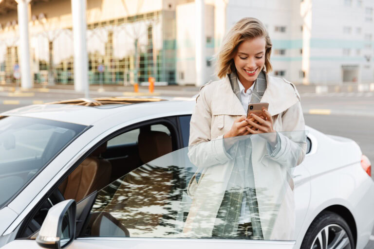Image of young businesslike woman standing by car and using cellphone outdoors